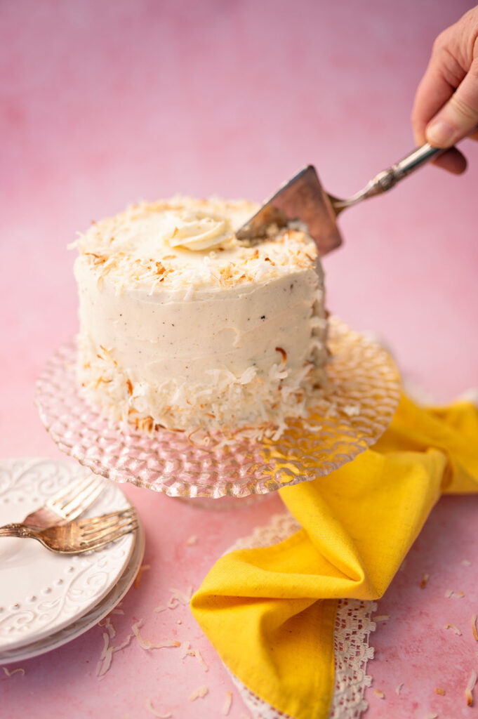 a hand is slicing a layer cake on a pink background with yellow linen napkin