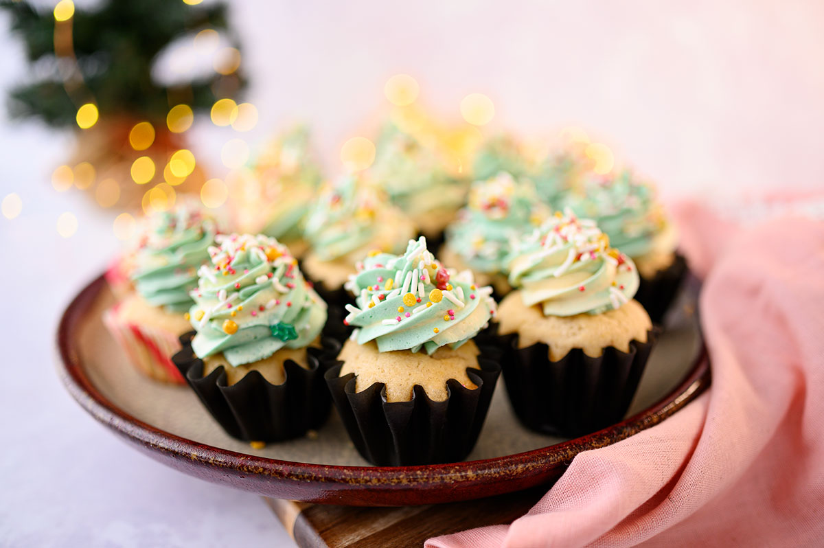 tray of a dozen piped cupcakes with sprinkles in front of a small Christmas Tree with twinkling lights