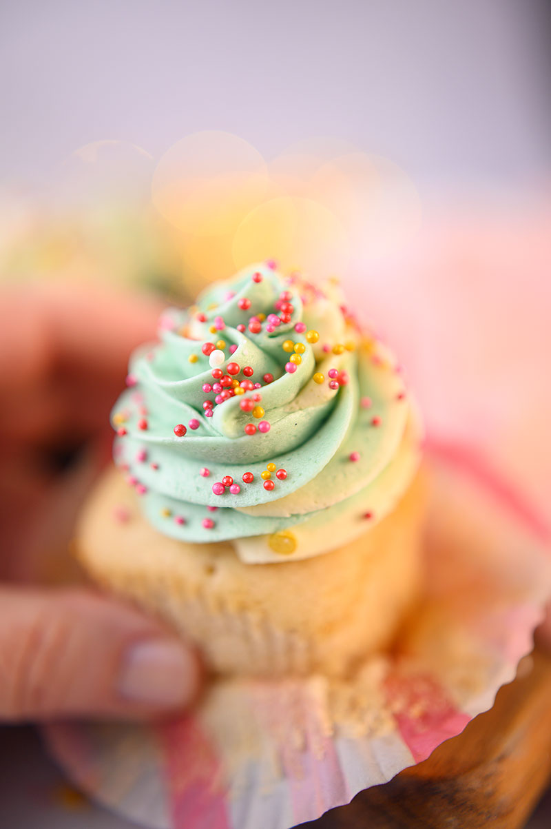 Close up of a hand holding a piped cupcake with light green frosting and pink and red sprinkles.