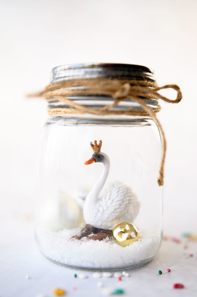 a white swan wearing a crown inside a mason jar with fake snow and jingle bells