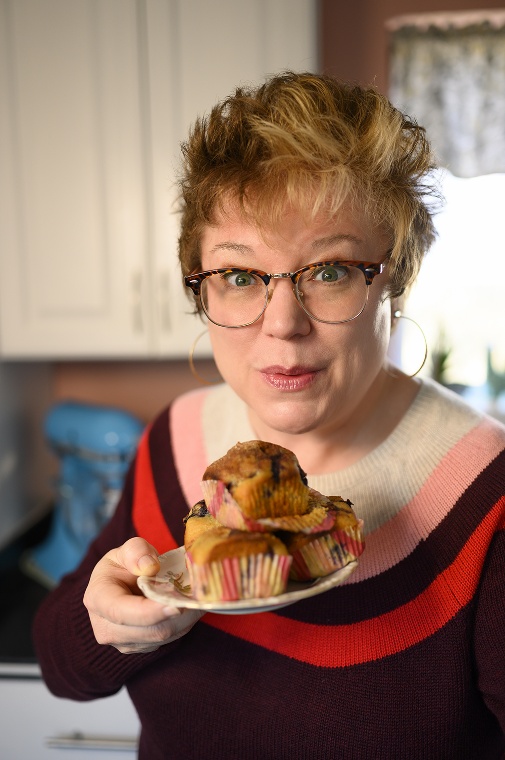 woman holding a plate of blueberry muffins in a home kitchen