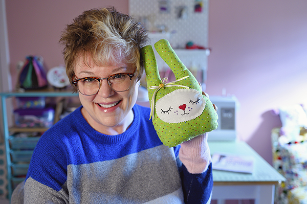 Woman holds a green bunny toy, handmade with her sewing machine also pictured.