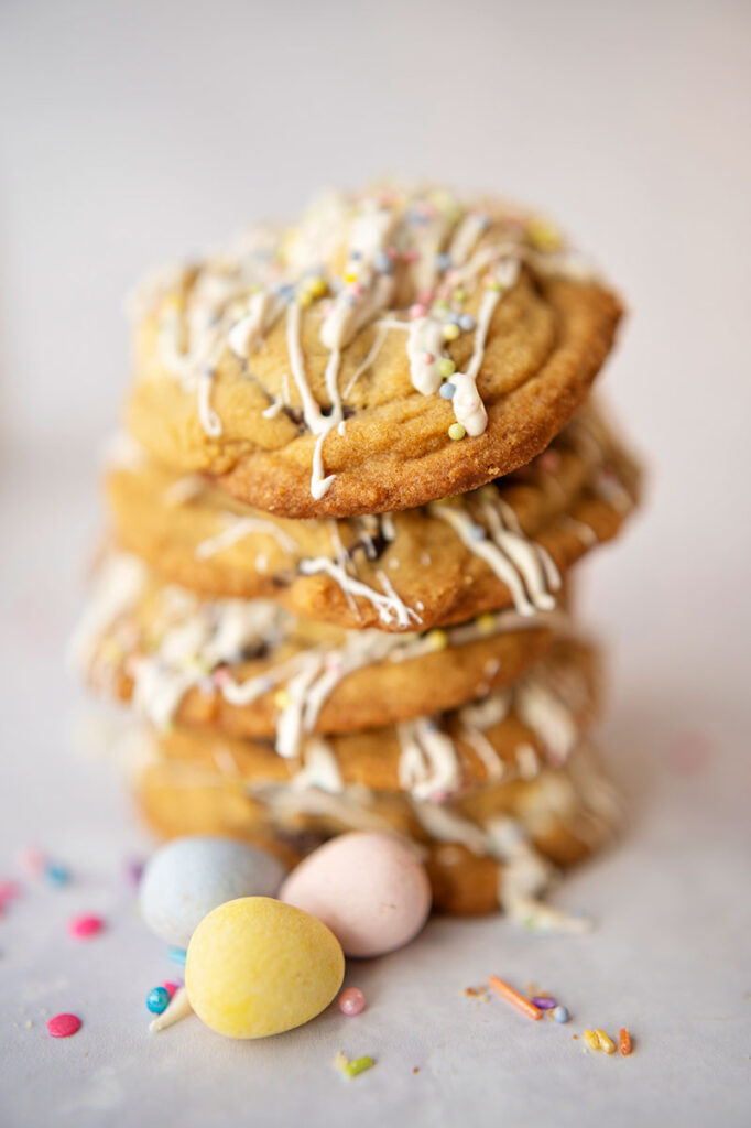 tall stack of cookies covered in white chocolate drizzle and sitting with mini candy coated pastel Easter eggs and colorful sprinkles