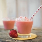 A glass of pink strawberry daiquiri is topped with whipped cream and a pink polka dotted straw. There is a fresh whole strawberry next to the glass for garnish.