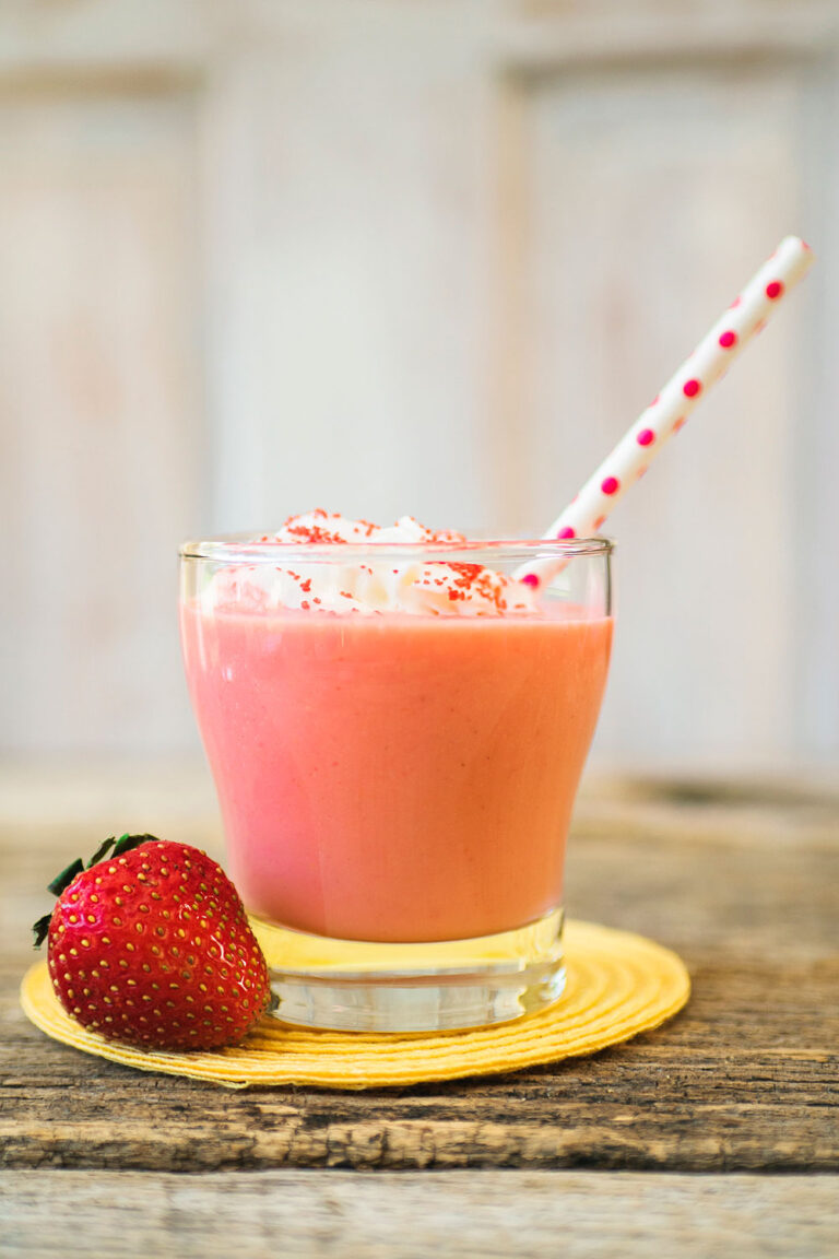 A glass of pink strawberry daiquiri is topped with whipped cream and a pink polka dotted straw. There is a fresh whole strawberry next to the glass for garnish.