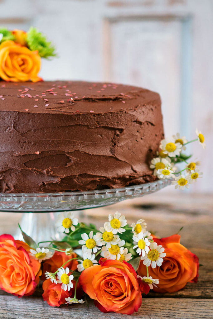 a large chocolate layer cake sits on a glass pedestal cake stand decorated with small white daisies and orange roses.