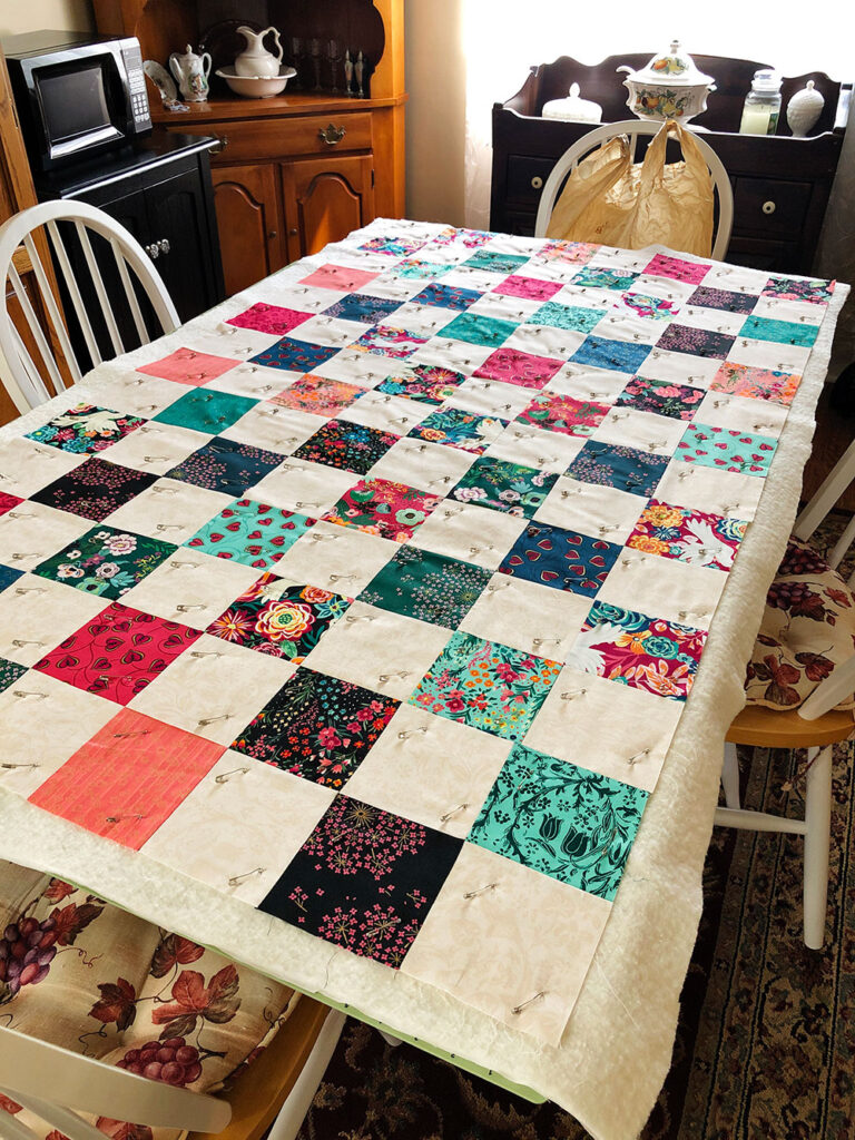 A patchwork quilt top is laid out flat on a table pin basted to the batting and backing.