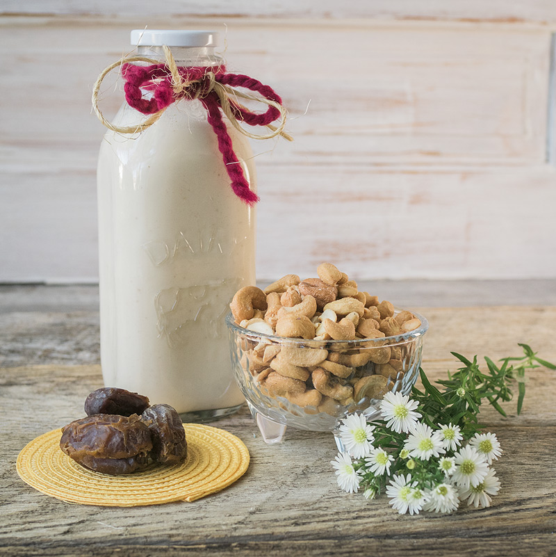 a bottle of chilled cashew milk sits with the ingredients used to make it, roasted cashews and medjool dates