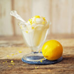 A vintage ice cream sundae dish is filled with lemon frozen yogurt and sprinkled with lemon zest. A whole fresh lemon sits next to the dish.