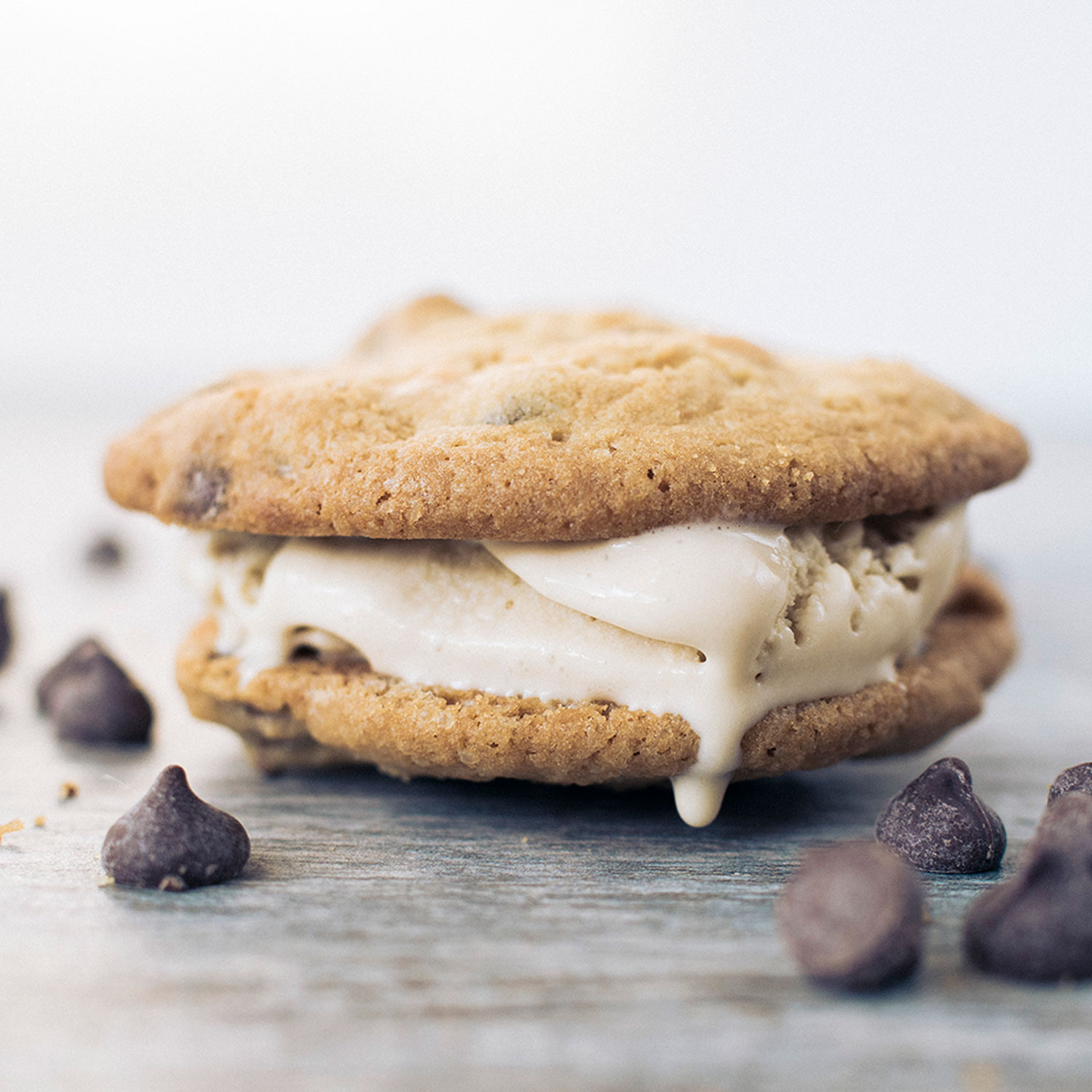 closeup of two chocolate chips cookies sandwiching vanilla ice cream that is slightly melting. Chocolate chips are scattered around