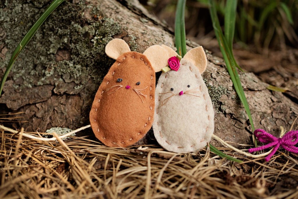 Two hand stitched felt craft mice posed together in pine straw at the base of a tree