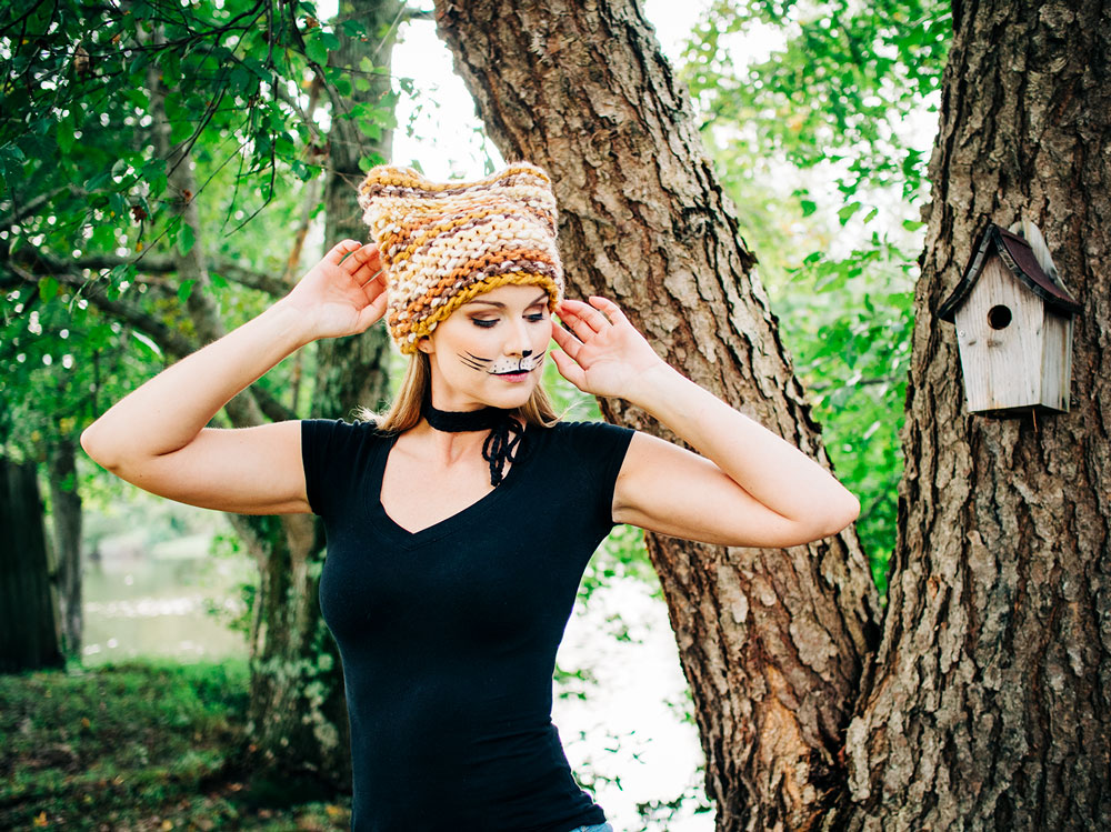 beautiful blonde model wearing makeup to look like a cat in a brown and golden chunky knit hat with ears