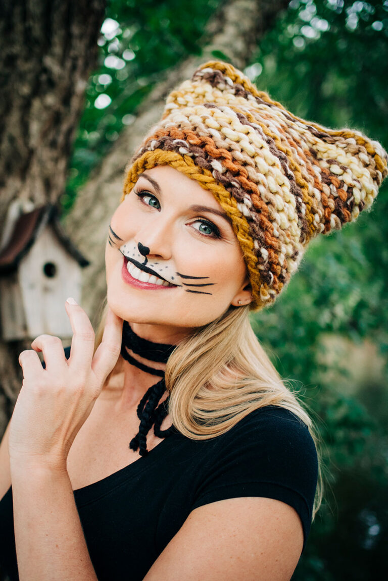 Smiling womans wears a multi colored knitted cat hat with cat makeup