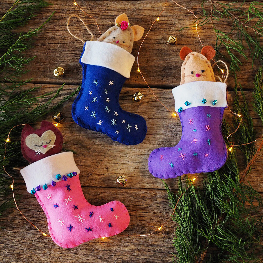 overhead view of three hand sewn felt stockings with 2 mice and a cat felt toys posed on barn wood with greenery and fairy lights