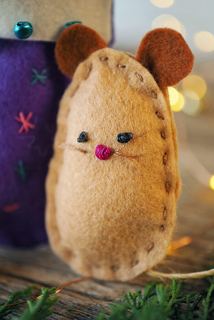 close up on a hand stitched felt mouse with fairy lights in the background and next to a hand stitched Christmas stocking