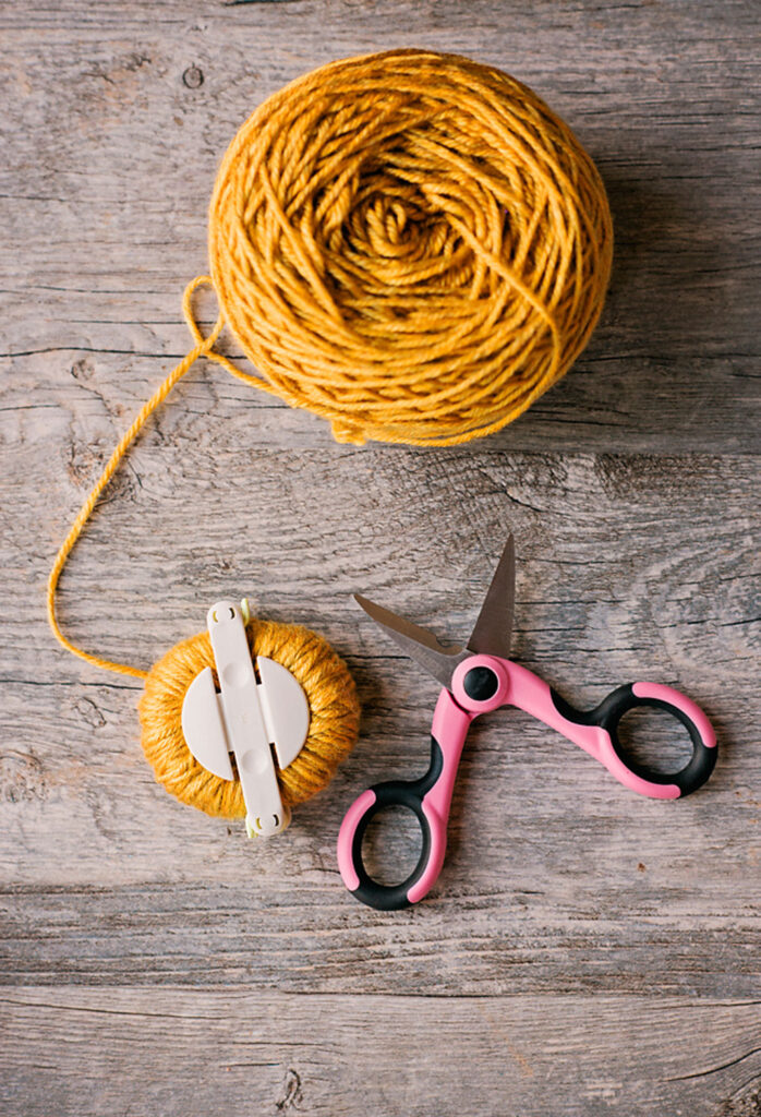 golden yellow yarn with a medium pom pom maker posed with pink scissors