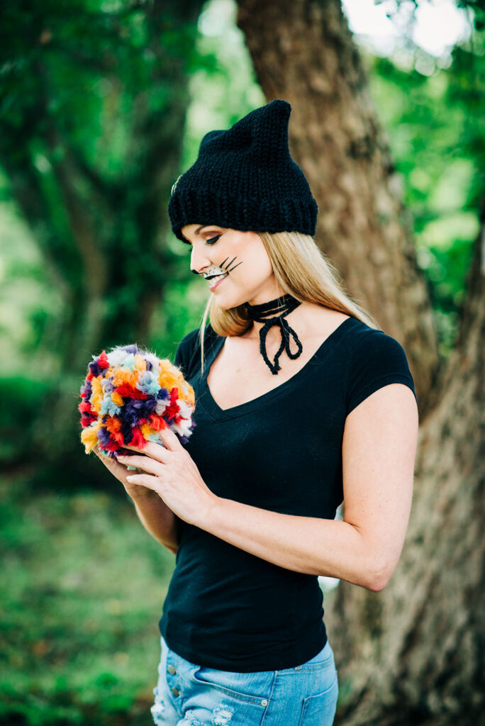 A blonde model in a black hat with cat makeup is holding a multi color jumbo pom pom