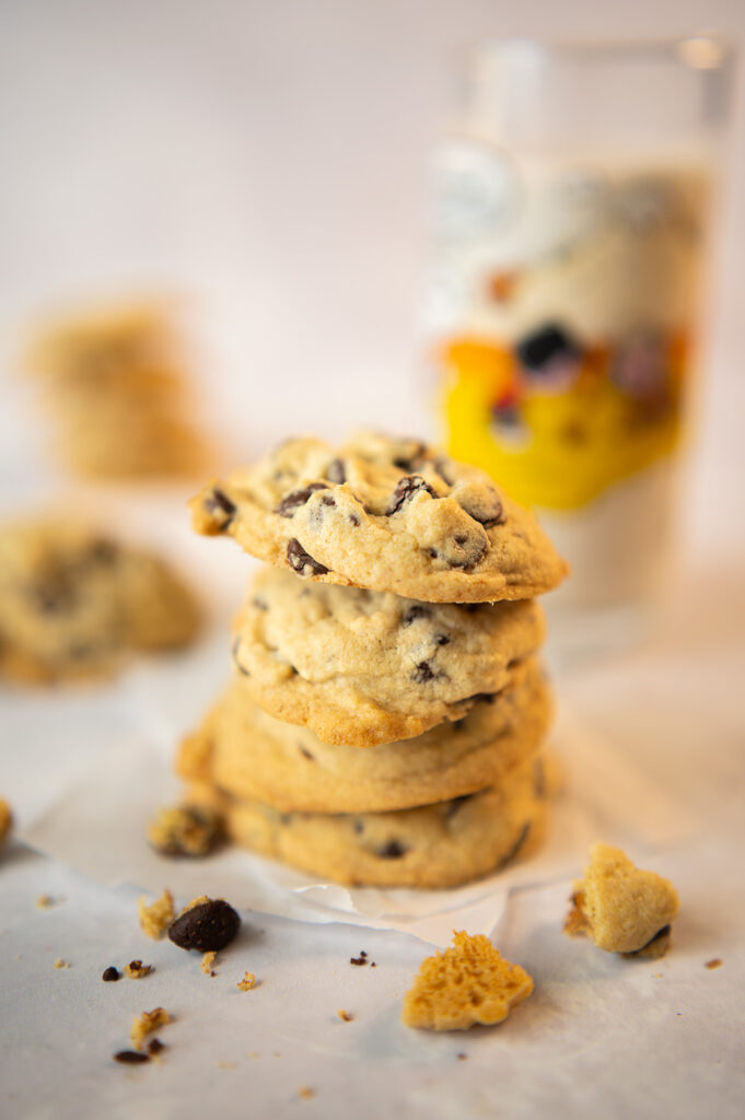 cookie stack closeup of thick, soft bake chocolate chip cookies. There is a glass of milk in the background and scattered crumbs of cookies in the foreground