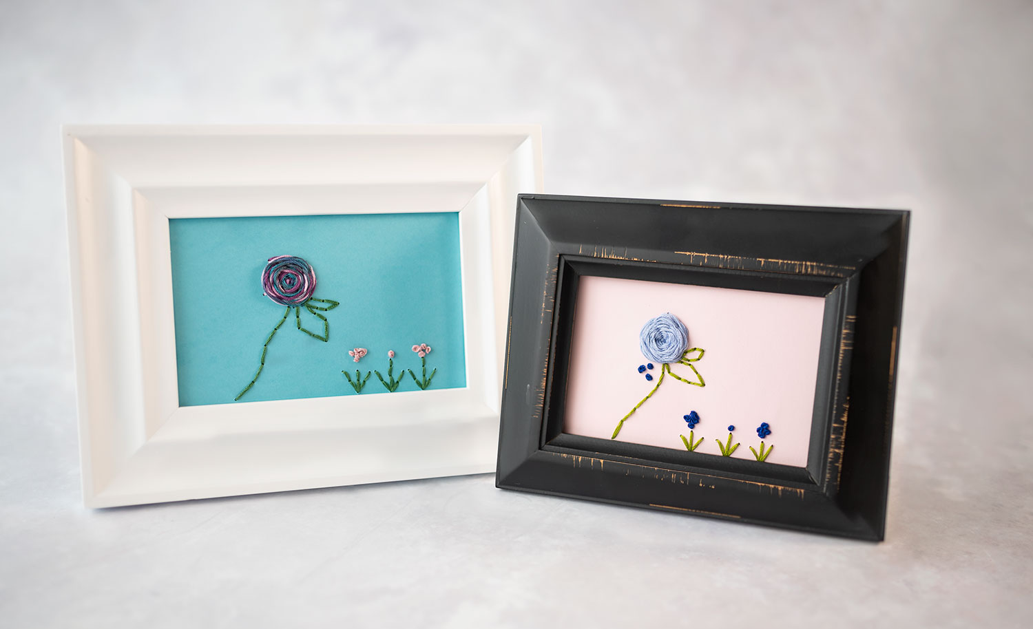 two framed, hand stitched flower designs. One in a white frame and one in a black frame. Both are hand stitched using the included pattern.