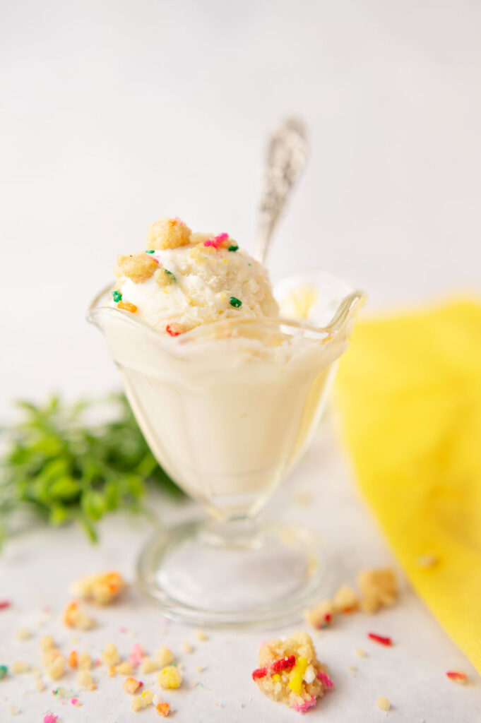 vintage glass dish with vanilla ice cream topped with multi colored toppings with yellow linen napkin and antique spoon