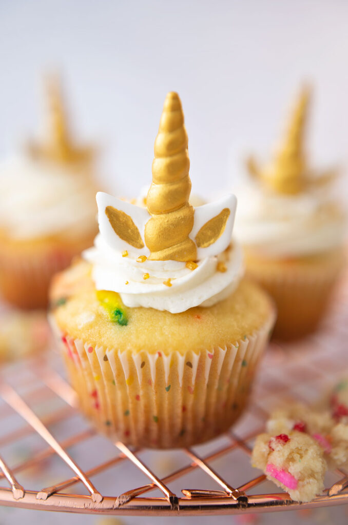 closeup of a vanilla cupcake with a polka dot wrapper and toppe with cloud white frosting and unicorn horn and ears