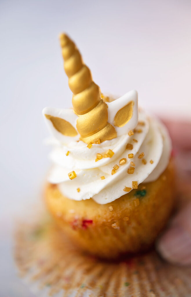 closeup view of a funfetti cupcake in multi color polka dot cupcake wrappers and topped with snow white frosting and a unicorn decoration favor. The wrapper is peeled away and a hand is holding the cake