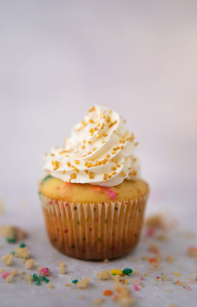 vanilla cupcake in a polka dot wrapper with a high swirl of snow white frosting and gold sanding sugar sprinkles