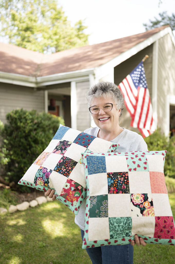 Smiling woman holds two large patchwork pillows in her front yard
