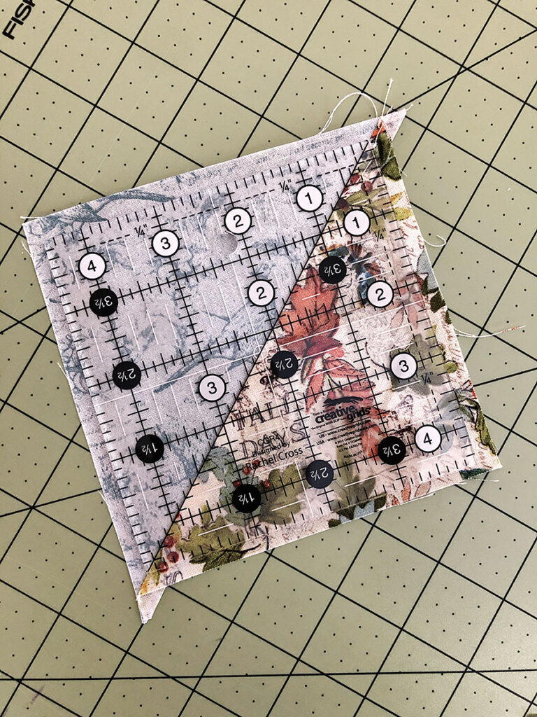 a square quilting ruler laying on top of a half square triangle block ready for trimming