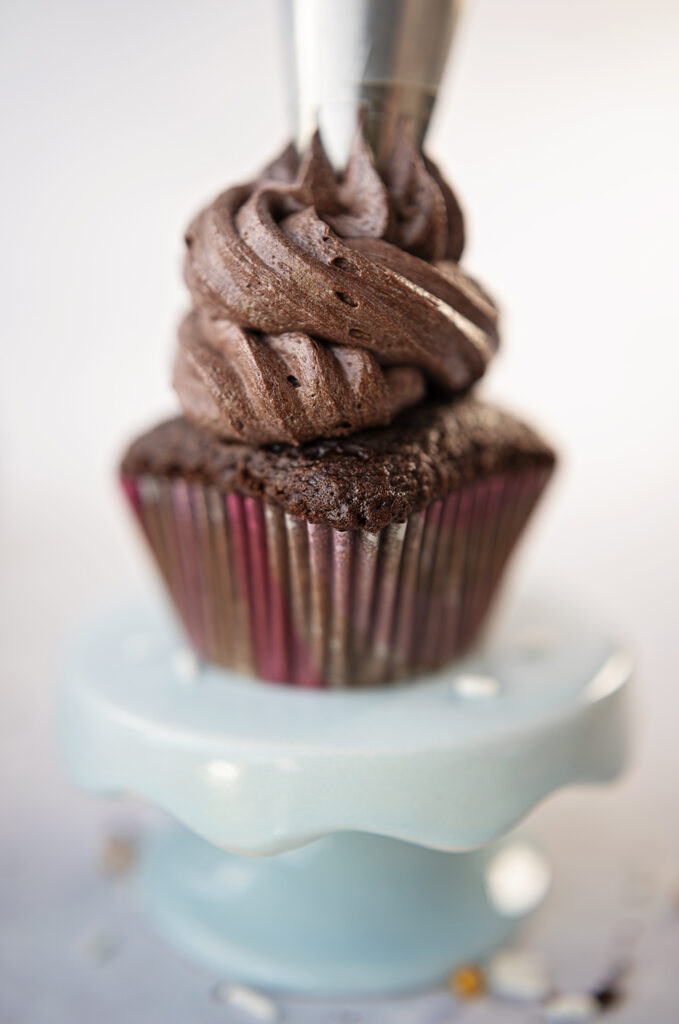 close up view of piping a high swirl of chocolate frosting on a chocolate cupcake