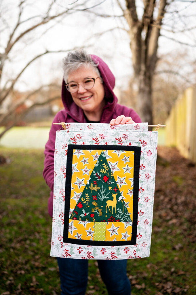 smiling woman holds a complete Christmas tree quilt block sewn into a mini quilt