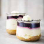 closeup of 2 small glass jar reveals 3 layers of dessert. A graham cracker base, creamy cheescake and a blue berry topping