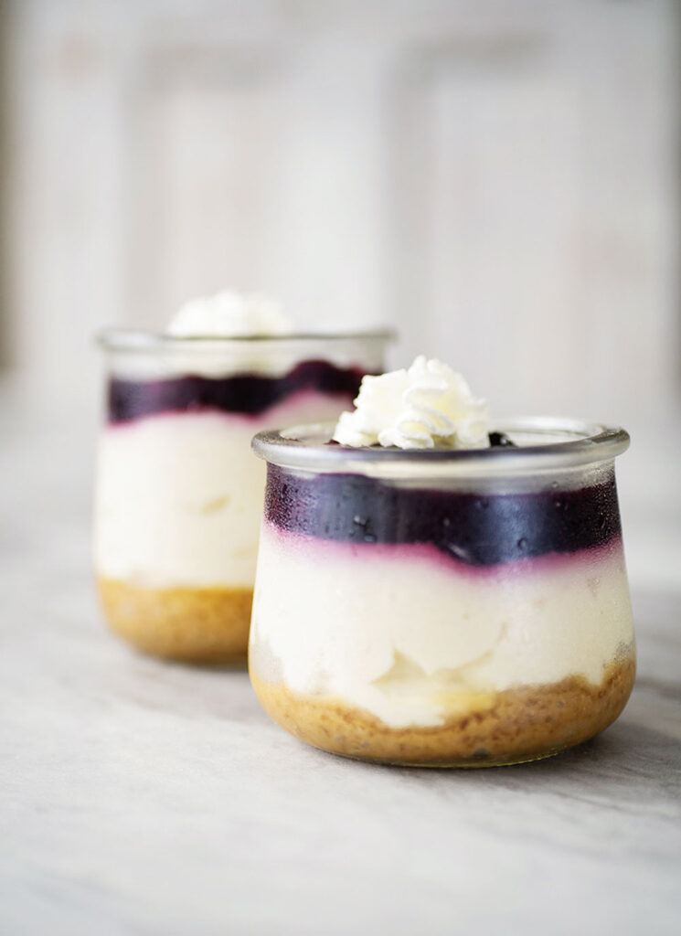  closeup of 2 small glass jar reveals 3 layers of dessert. A graham cracker base, creamy cheescake and a blue berry topping