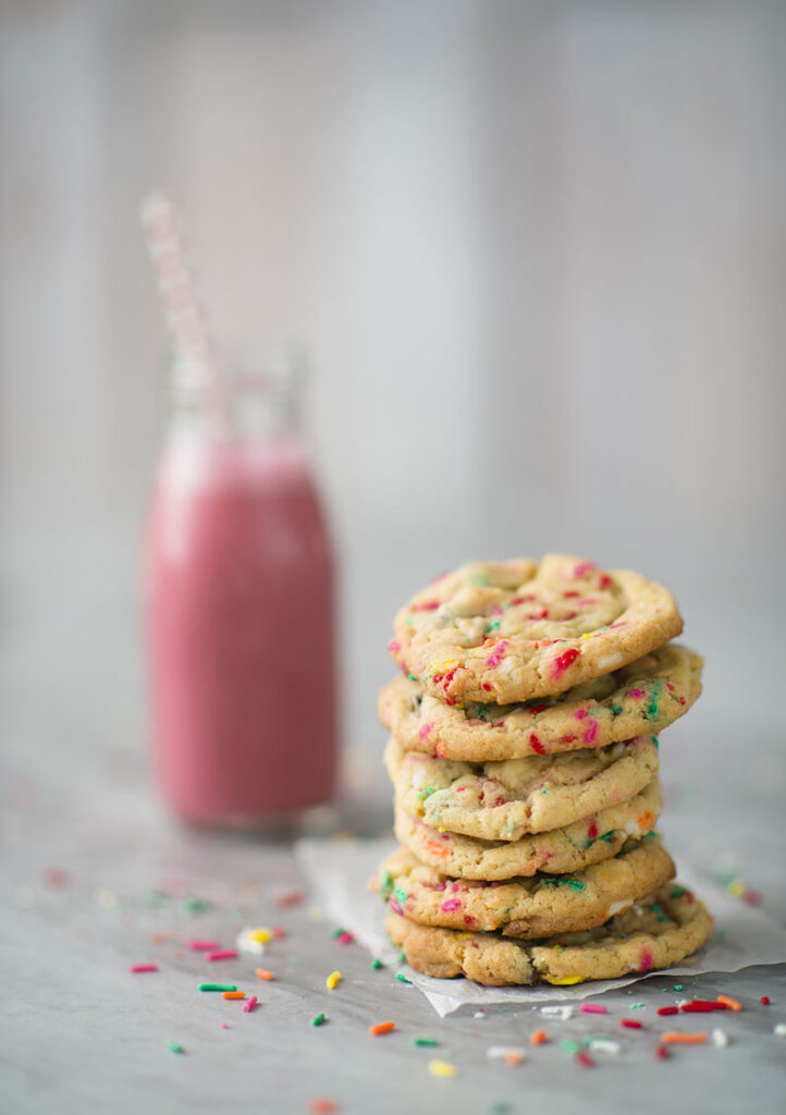 stack of colorful cookies filled with sprinkles, sprinkles on the table top and a pink strawberry milk