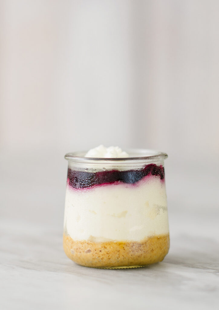 a small glass jar reveals 3 layers of dessert. A graham cracker base, creamy cheescake and a blue berry topping