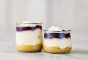 2 small glass jar reveals 3 layers of dessert. A graham cracker base, creamy cheescake and a blue berry topping