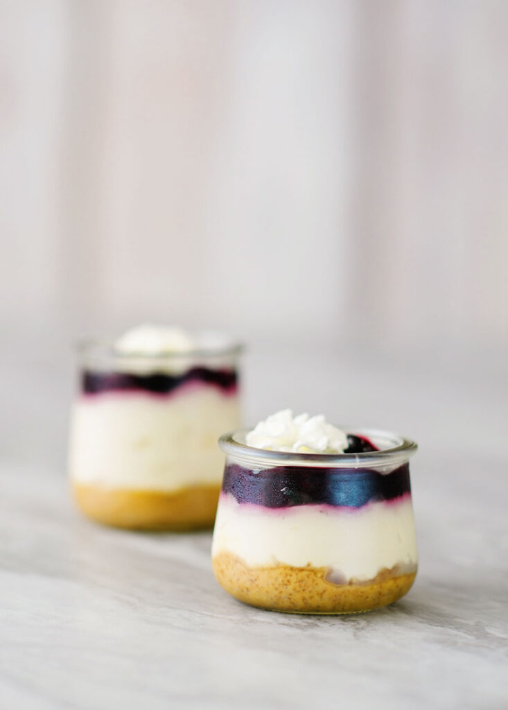 2 small glass jars reveals 3 layers of dessert. A graham cracker base, creamy cheescake and a blue berry topping