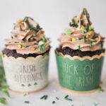 Two chocolate cupcakes in Irish themed cupcake wrappers piped with mile high mocha frosting and shamrock sprinkles