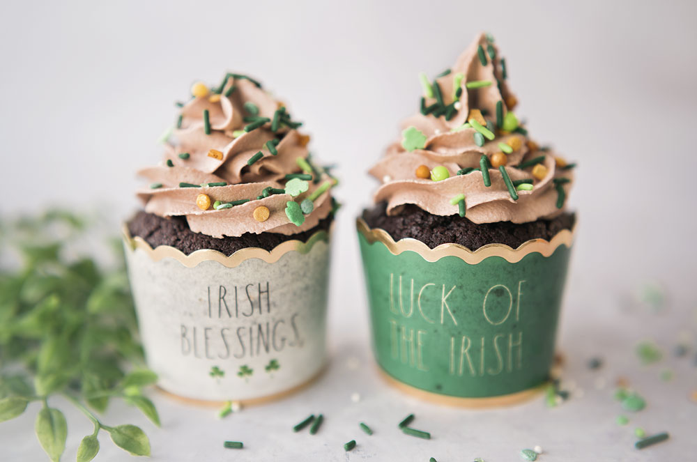 Two chocolate cupcakes in Irish themed cupcake wrappers piped with mile high mocha frosting and shamrock sprinkles