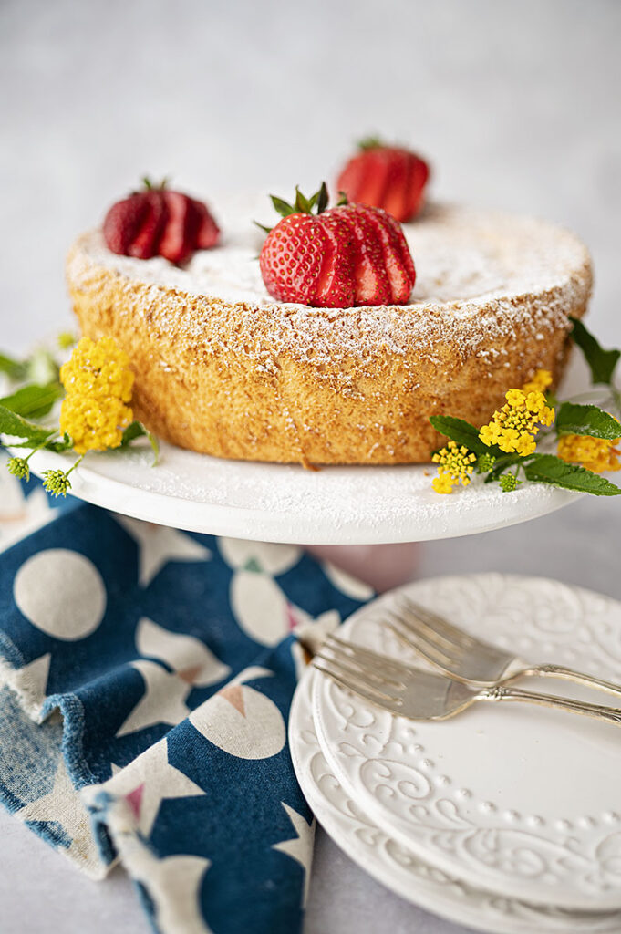 An angel food cake on a cake plate topped with fresh sliced strawberries and decorated with delicate yellow flowers