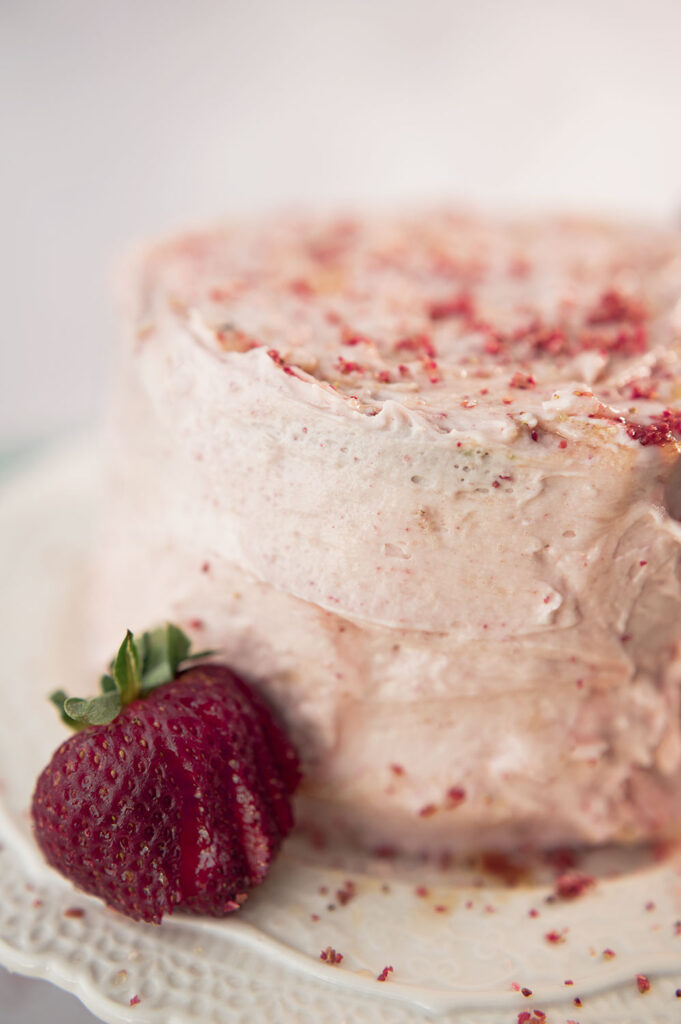 a pink frosted cake on a lacy white cake plate with fresh strawberries