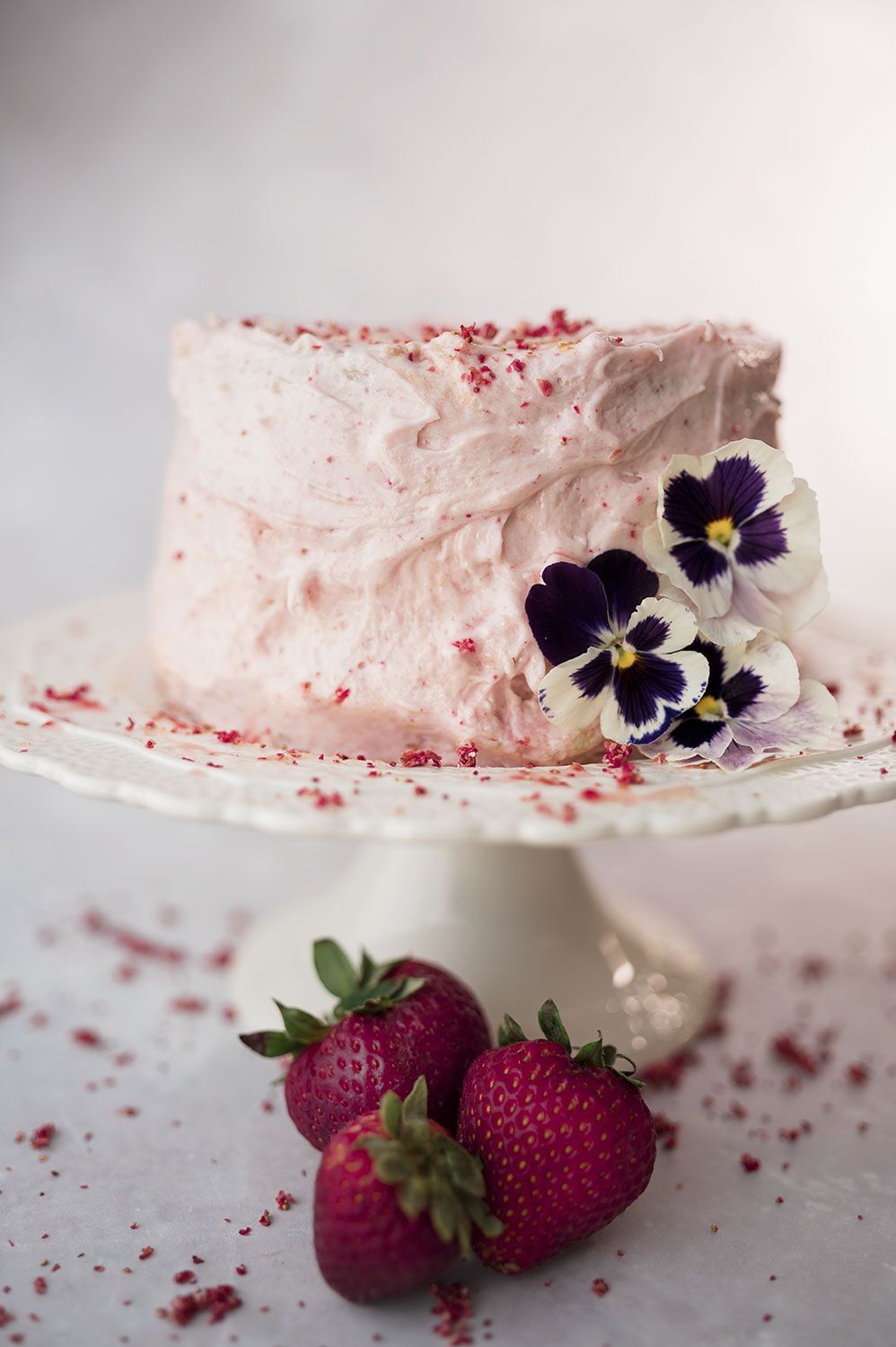 a pink frosted cake on a lacy white cake plate with fresh strawberries