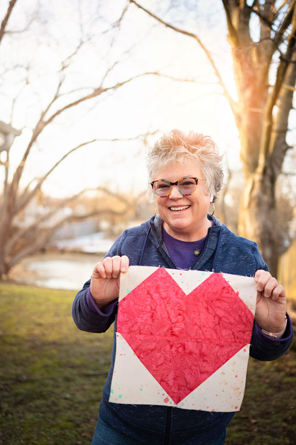 Smiling woman holds a pieced quilt block of a pink heart