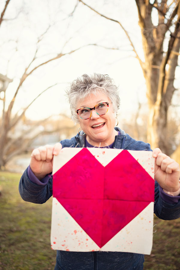 Smiling woman holds a pieced quilt block of a pink heart