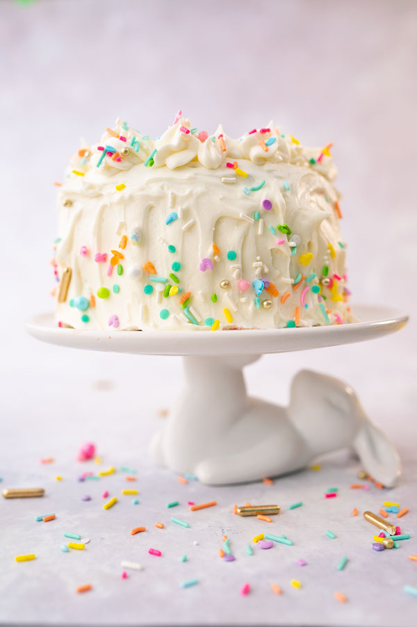A white frosted cake decorated with colorful sprinkles on a cake plate that looks like a bunny