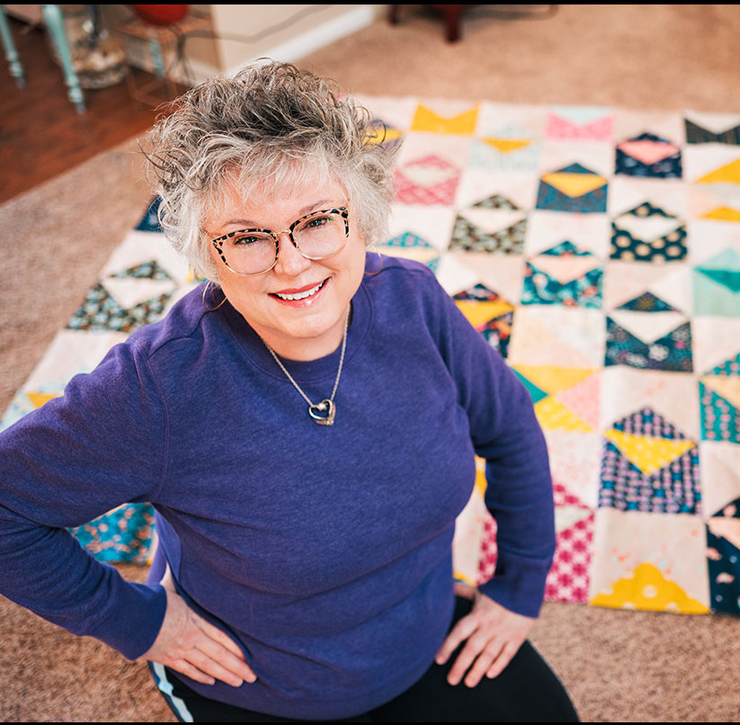 A smiling woman in a purple sweat shirt sits in front of a large quilt