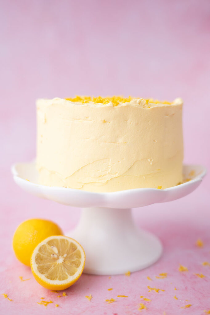 An elegant looking lemon cake frosted in pastel yellow buttercream on a white cake stand